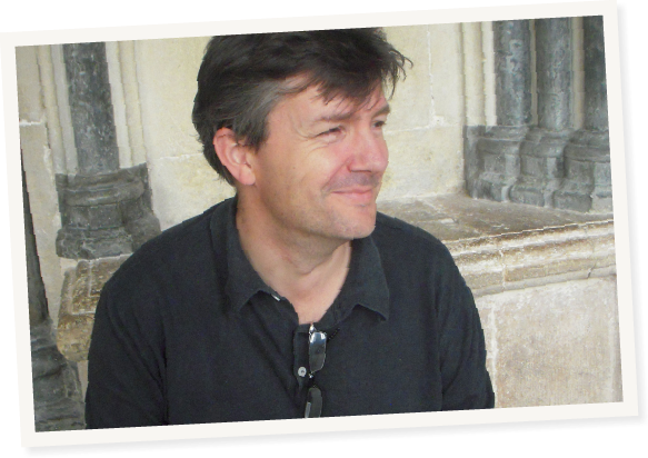 Pete, smiling, sitting in a black shirt in a grand looking stone building. He has sunglasses hanging on the top undone button of his polo shirt and he's looking off to the image right.
