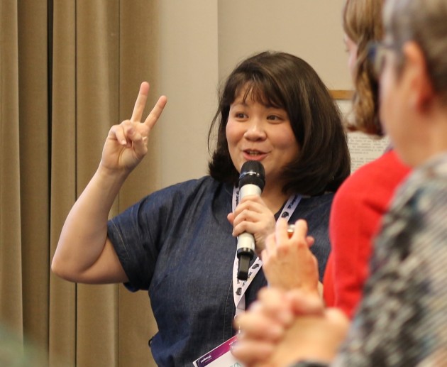 photo of Clara presenting into a handheld microphone, gesturing in the air, people around her listening.