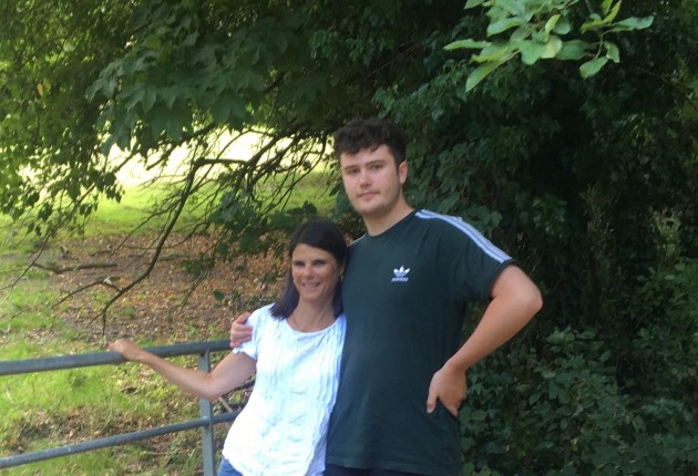 Helena wearing a white t-shirt stands at a farm gate with her son Matthew wearing a dark blue sports top, with a field and trees behind