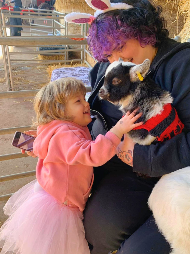 A CHILD AND MOTHER ENJOYING CUDDLING A BABY GOAT WEARING A JUMPER