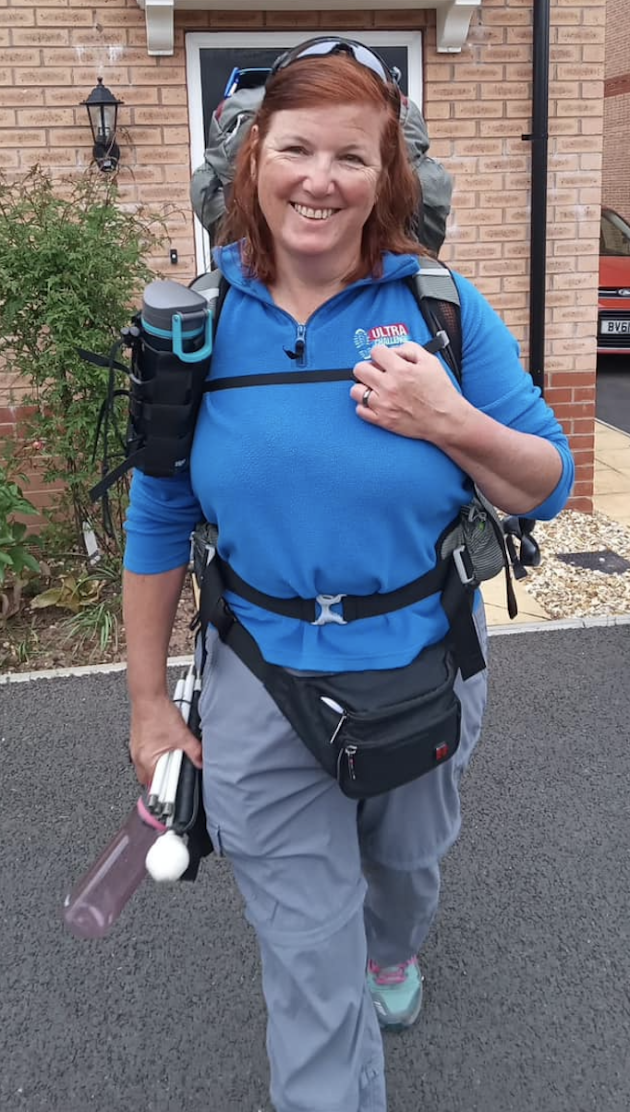 Photo of Nicky Button standing in front of a house ready to go walking. She is wearing a blue shirt, grey trekking trousers and has a large backpack and water bottles.