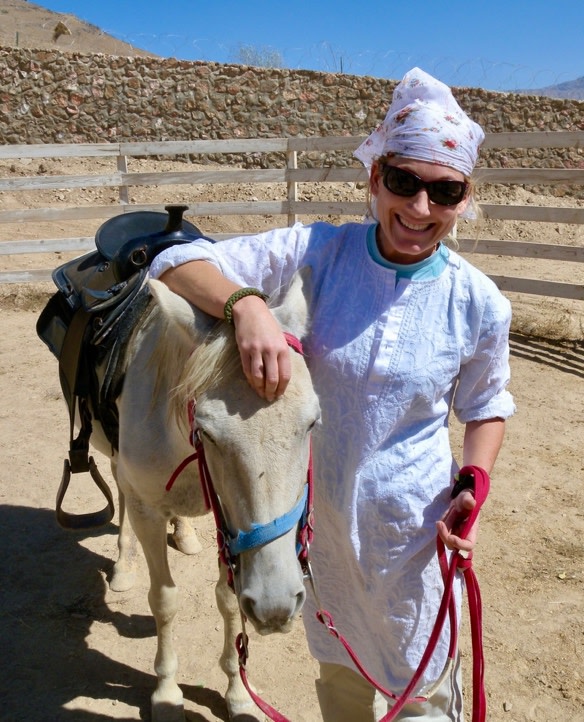 Holly is pictured wearing a head scarf and sunglasses in a desert location stranding with her arm resting on a horse.