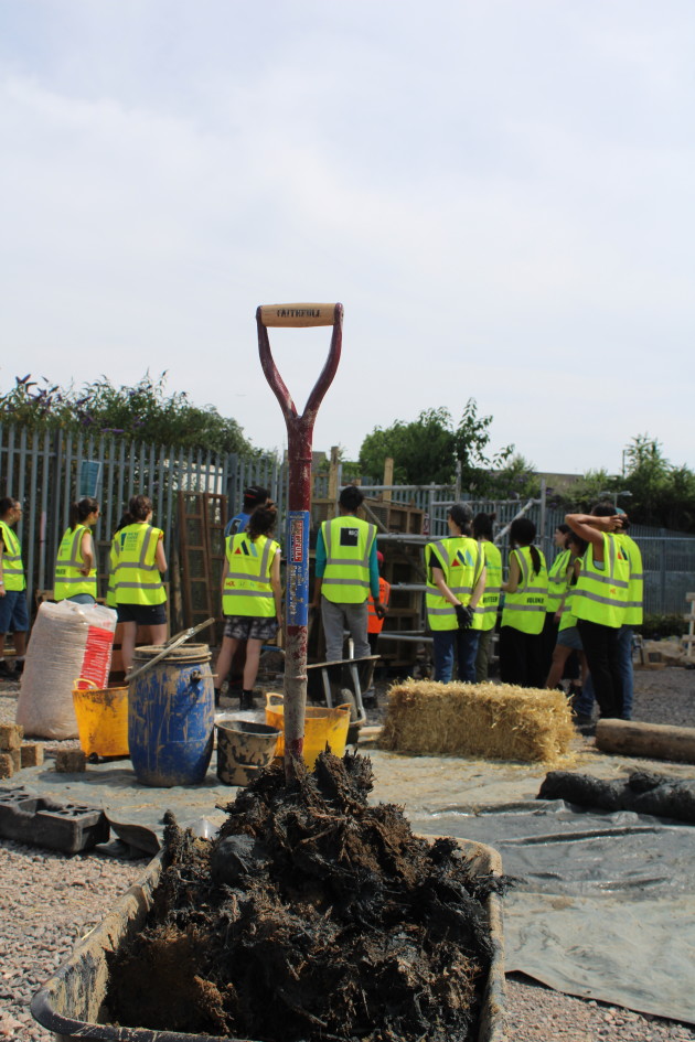 A group of people gather looking at a formwork structure. In the foreground is a spade stuck into a pile of cob.