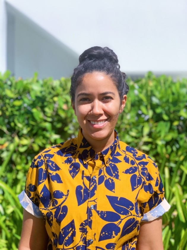 Kristie, who has tan skin and brown hair that is pushed back into a bun, smiles at the camera. Kristie is wearing an orange button down with blue leaves and is standing in front of a bush.