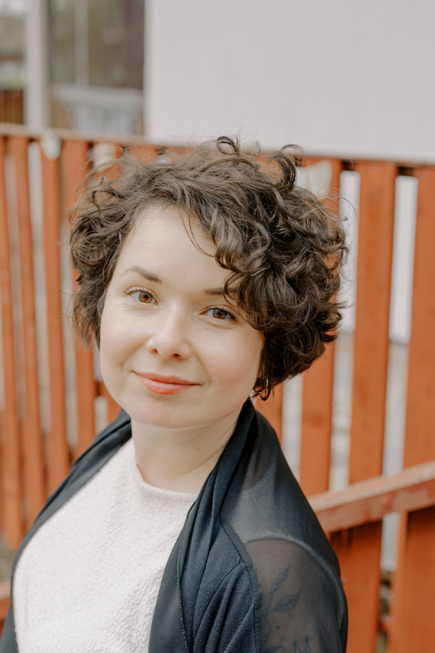 Zula Rabikowska headshot image. White smiling woman with curly brown hair and a white top against an orange fence.