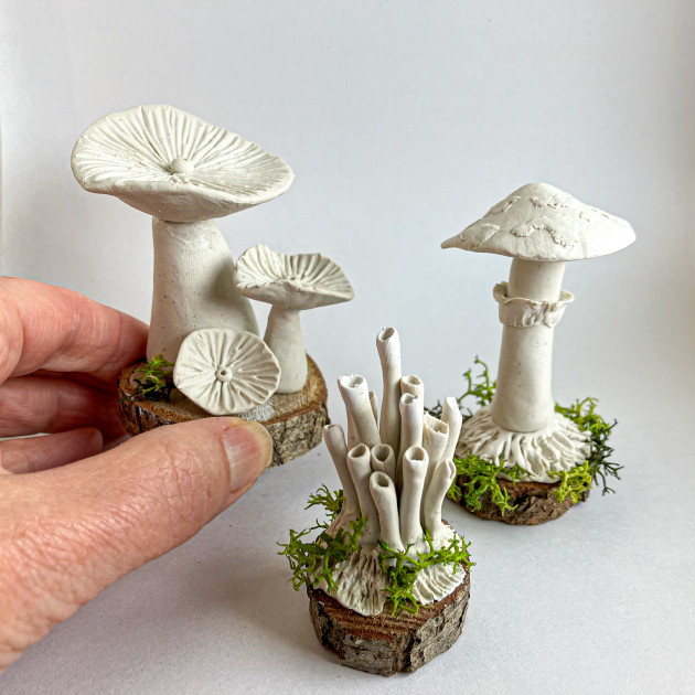 Three small white sculptures of fungi. One of them is being held in Kathryn's hand. They each stand on a little wooden base.